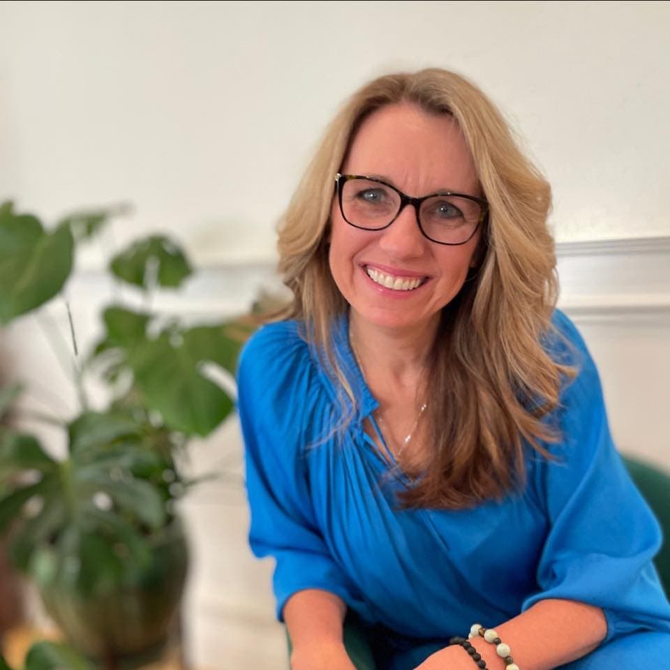 Joanna, founder of Sparkle of Light Wellness, smiling warmly. She has shoulder-length blonde hair and glasses and is wearing a brown blouse.