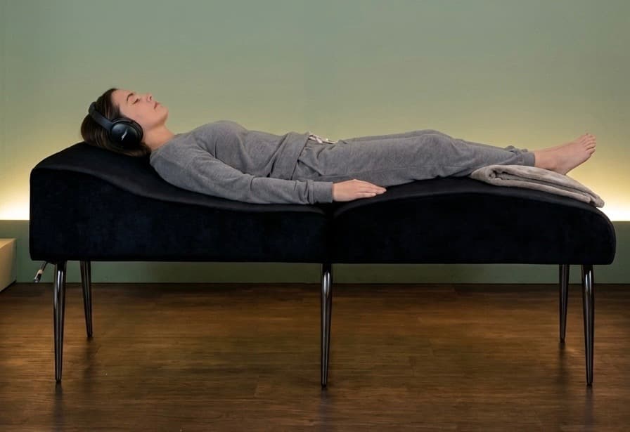 A person reclining on the Sound Lounge's wooden lounger, wearing headphones with their eyes closed.
